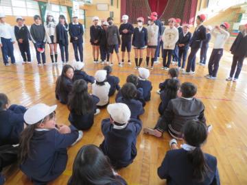 体育館で、立って屋内スポーツクラブの紹介をする6年生とその話を床に座って聞く3年生たち