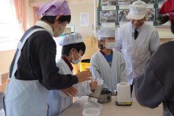 エプロンと頭巾をかぶった児童と教師が豆乳を鍋に移しているようす