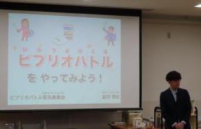 モニターに表示された文字「ビブリオバトルをやってみよう」と、その隣に立って話す男性