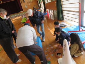 体育館で児童たちにめんこの手本を見せる長寿会の皆さんの様子