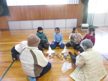 児童たちと老人会の方が体育館の床に座り、昔の遊びを見たり聞いたりしています