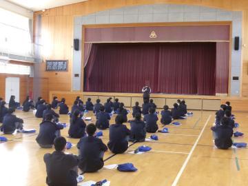 体育館のステージ前中央に教員が立ち、話をしています。生徒は整列し、体操座りで前を向いて教員の話を聞いています。