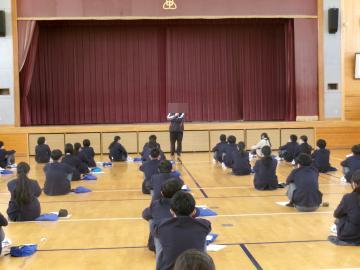 体育館のステージ前中央に教員が立ち、話をしています。生徒はステージ前に整列し、顔をしっかりあげて教員の話を聞いています。