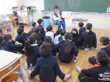 絵本の読み聞かせを教室の床に座ってたのしむ2年生
