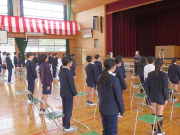 体育館の並んだパイプ椅子の前で起立する生徒たち