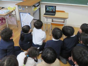 教室で座り、オンラインの絵本読み聞かせをパソコンの前に座って見る児童たちの様子
