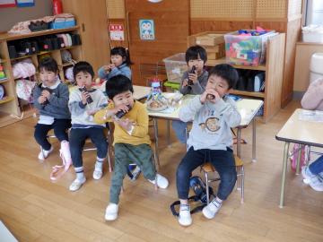 教室で椅子に座って、みんな同じ方向をむいて恵方巻を食べる幼児