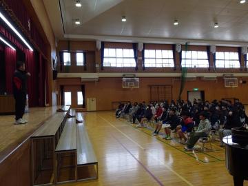 体育館で壇上に立つ男性教師と、椅子に座って話を聞く6年生のようす