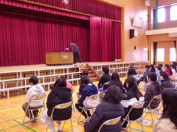 体育館の壇上で卒業証書のもらい方を示す男性教師と、椅子に座って話を聞く6年生のようす