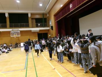 体育館の壇上の前で6年生が4年生から大学帽を受け取っている