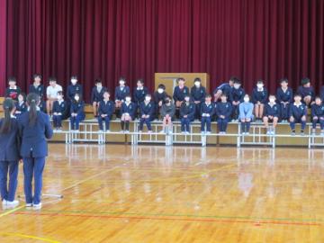 体育館で5年生によるはじめのことばを聞く6年生の様子