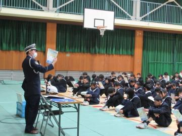 体育館で、チラシを見せて説明する警察官と床に座ってチラシを見る生徒達