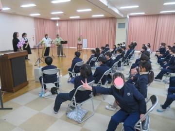 公民館でサックス演奏をする方と、それを椅子に座って聞く子供たちのようす