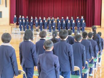体育館の壇上に整列する6年生と自席で起立している5年生