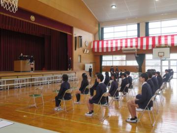 体育館の壇上で卒業証書を受け取る生徒と教師と、椅子に座っているほかの生徒たち