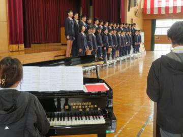 体育館の壇上のひな壇で整列して歌う6年生とピアノを演奏する教師のようす