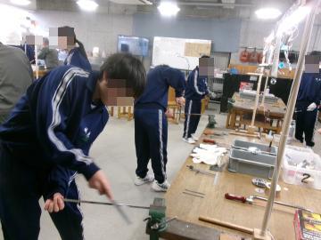 技術室で鉄の棒をたたいてキャンドルスタンドの作成をする生徒達