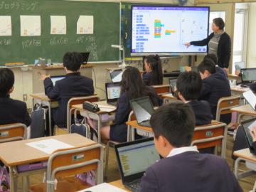 教室で席に座る生徒達にモニターを使ってプログラムの説明をする校長先生