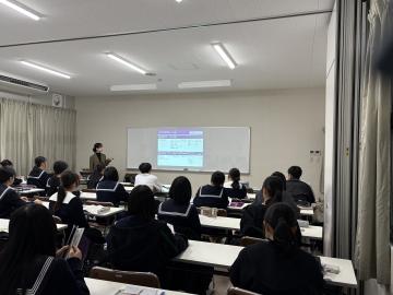 会議室でスクリーンに資料を映しながら説明する講師と、椅子に座って話を聞く生徒達1