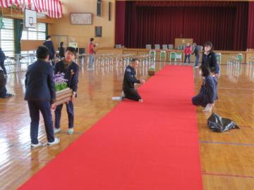 体育館のレッドカーペットの両脇に桜草のプランターを運ぶ5年生