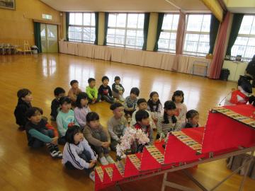 遊戯室に飾ってあるひな人形の前に子供たちが集まって座っている写真