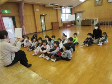 遊戯室でステージの段差に腰かけて紙芝居を読む先生と床に座っている園児たちの様子