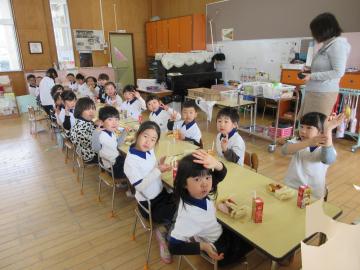 教室の長いテーブルに着席しているこどもたちがカメラに向かってピースサインをする園児たちの様子