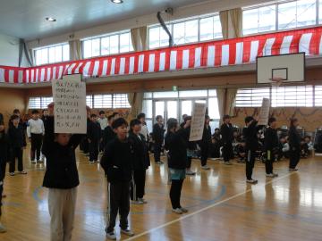 4年生が歌詞のかかれた画用紙を頭の上で掲げながら、立って歌うようす