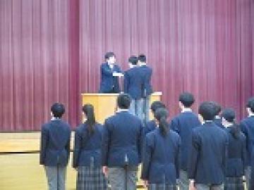 体育館の壇上で修了証を校長先生から受け取る2年生の代表生徒の様子