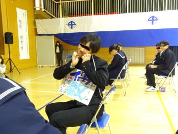 体育館で椅子に座る生徒達と、カメラに向かって握った手を頬の横においてポーズをする生徒