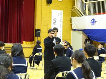 体育館で椅子に座っている生徒達の前に立ってマイクで話す3年生のようす