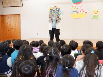 教室で立って紙芝居を読む先生と、その紙芝居を座って見る幼児たち