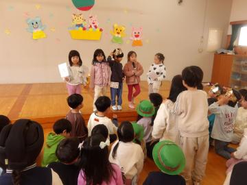 遊戯室のステージに立って話す子供たちと、その様子を見る子供たち