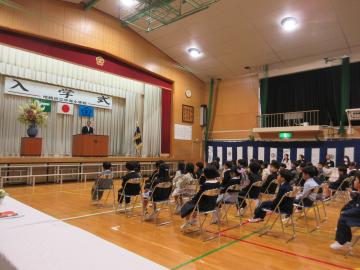 体育館の壇上に先生が立って、椅子に座った児童たちが整列して話を聞く様子