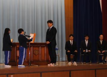 体育館の壇上で、離任する校長先生にメッセージを読む児童と花束を渡す児童