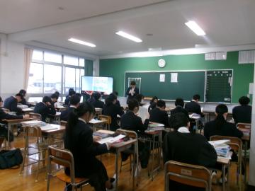 教室で教師が前に立ち黒板とスクリーンを使って生徒に配布資料の確認を行っている様子