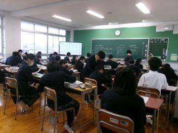 黒板のある教室で多数の生徒が座って机に向かい配布資料を見ている