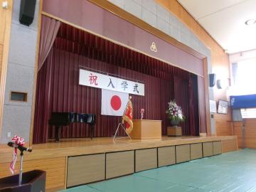 学校の体育館のステージに日の丸の国旗と祝入学式の横断幕が飾られ演台とピアノと花が配置されている光景