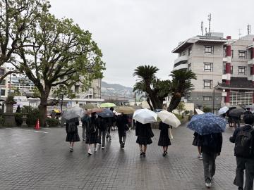 雨の街路を傘をさして生徒たちが歩く風景で背景に樹木と集合住宅が見える