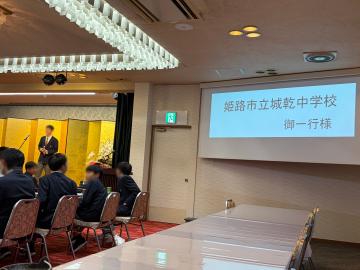 会議室でスクリーンに「姫路市立城乾中学校 御一行様」と表示され出席者が前方を向いて座り演壇で話す人がいる様子