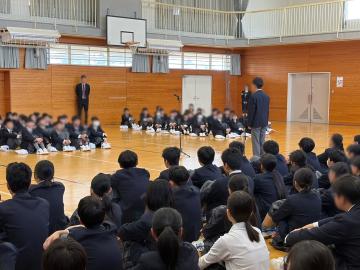 体育館の床に座って列になった一年生を前に生徒代表が立って話している対面式の様子