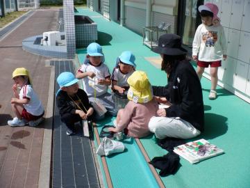 首からミニ図鑑を下げた子供たちが、飼育ケースに入っている虫を見ている様子