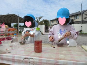 青い帽子をかぶった二人の子どもが屋外のテーブルで水や容器を使って遊んでいる様子