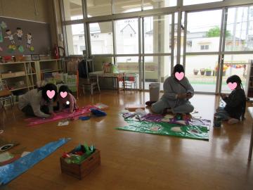 広い教室で床に敷いたシートの上に座って遊ぶ数人の園児たちと先生が鯉のぼりを作っている様子