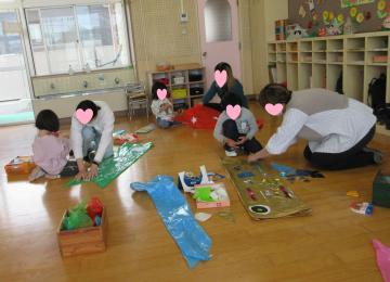 部屋の床に座って先生と子どもたちが遊具や工作材料で遊んでいる様子