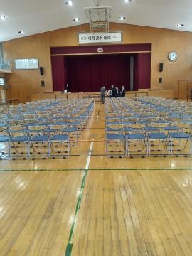 体育館のフロアに整然と並んだ折りたたみ椅子の列と舞台の赤いカーテンを背景に数人が舞台前に立っている