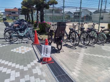 駐輪場のフェンス沿いに並んだ複数の自転車と手前に置かれた赤いコーンと注意看板がある屋外の風景