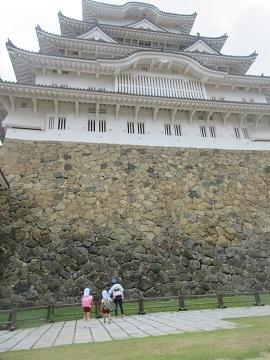 白い層状の天守が大きな石垣の上にそびえ立ち、手前に3人の園児たちが歩道に立っている様子