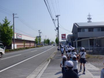 白い帽子をかぶった子どもたちが建物や電柱が並ぶ通学路の歩道を列になって歩いている風景
