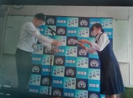 二人の人物が屋内で青と白のロゴ入り背景の前で互いに書類を差し出しながら礼をしている